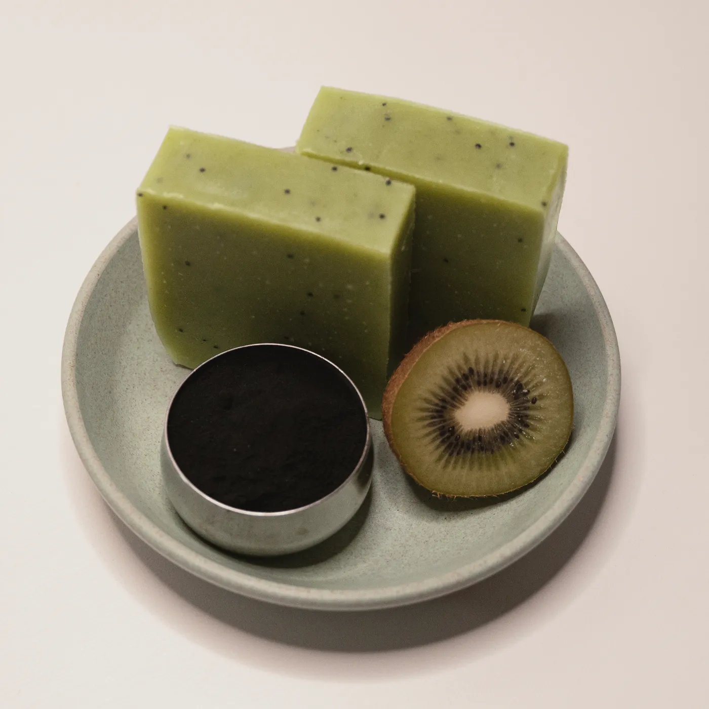 Two bars of Kiwi cucumber and spirulina soap on a green dish with spirulina powder and a halved kiwi