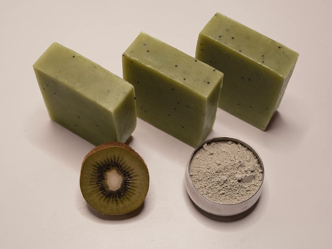 Three bars of Kiwi soap in a row with clay powder and a halved kiwi